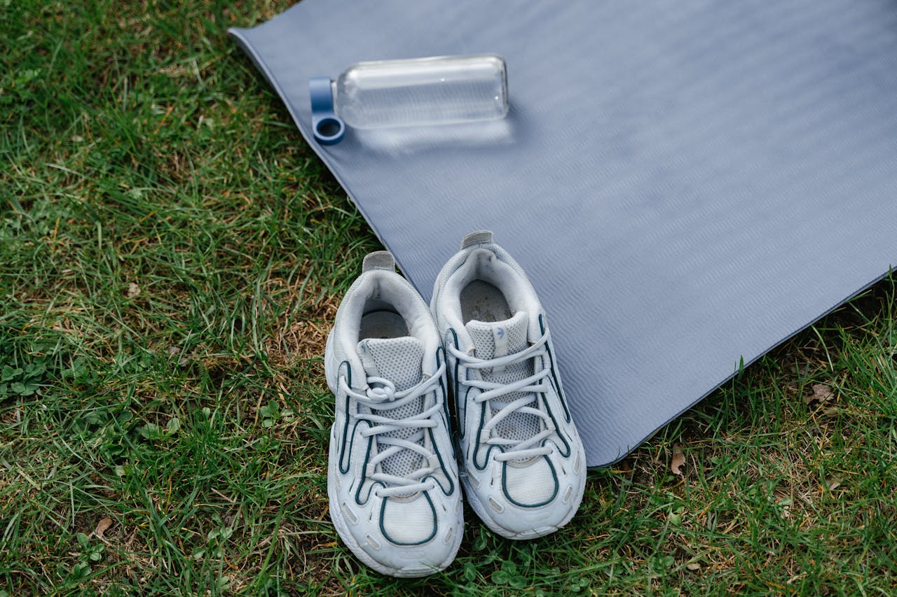 Pair of sneakers with a yoga mat and water bottle on grass, ideal for outdoor workouts.